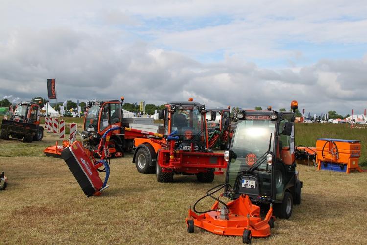 Pomarańczowe maszyny rolnicze Holder z różnym osprzętem na polu, w tle widoczne stoiska wystawowe i pochmurne niebo. Prezentacja sprzętu.