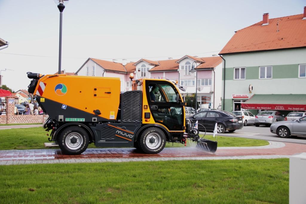 Żółto-czarna zamiatarka uliczna Muvo czyści chodnik na tle budynków i zaparkowanych aut. Woda spryskuje czyszczoną powierzchnię. Widok z boku, zieleń trawnika.