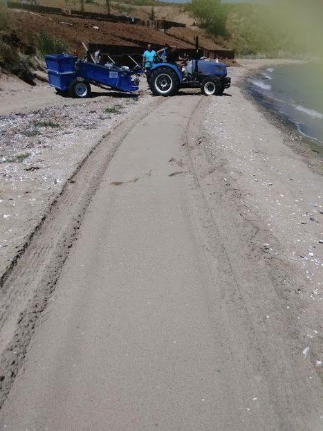 Niebieski traktor z przyczepą na piaszczystej plaży, ślady opon na piasku, w tle woda i brzeg. Praca maszyny na nabrzeżu.