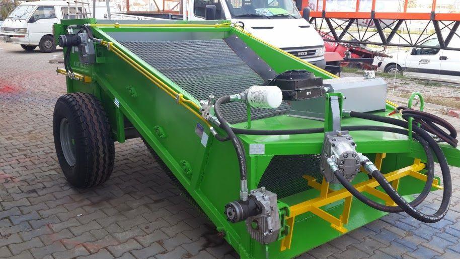 Zielona maszyna rolnicza z czarną siatką, widoczne opony, hydraulika i elementy konstrukcyjne na tle pojazdów. Sprzęt gotowy do pracy w polu.