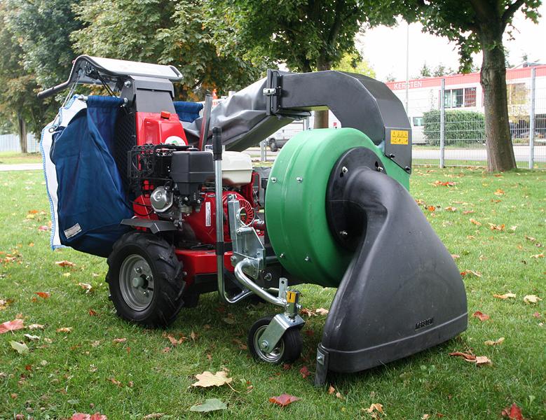 Samobieżna maszyna do zbierania liści na trawniku, z workiem i turbiną ssącą. Widoczne jesienne liście na trawie i budynek w tle.