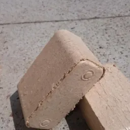 Dwa brykiety drzewne RUF z widocznymi otworami wentylacyjnymi, ułożone na betonowej posadzce w jasnym oświetleniu. Częściowo widoczny worek z brykietami.