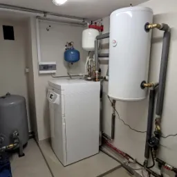 Kotłownia z kotłem gazowym, zbiornikiem CWU, naczyniami przeponowymi i rozdzielnicą elektryczną na ścianie, widoczne liczne rury i zawory.