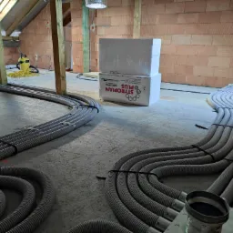 Szare, elastyczne rury wentylacyjne ułożone na betonowej podłodze poddasza w trakcie montażu, widoczne materiały izolacyjne i odkurzacz przemysłowy w tle