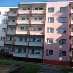 Wielopiętrowy budynek mieszkalny z balkonami, pomalowany na biało i różowo, z ciemnoczerwonymi akcentami, widziany z poziomu gruntu, z fragmentem placu zabaw i zaparkowanym vanem.