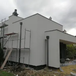 Świeżo otynkowana, biała elewacja nowoczesnego domu jednorodzinnego w trakcie wykańczania, widoczne rusztowania, zabezpieczenia i elementy wykończeniowe na dachu, pochmurne niebo w tle.