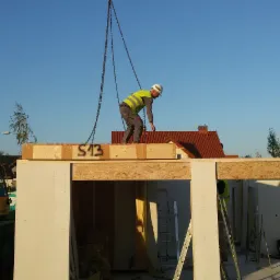 Montaż drewnianego stropu domu szkieletowego za pomocą dźwigu, pracownik w kasku i kamizelce odblaskowej na górze, widoczny numer S13 na belce.