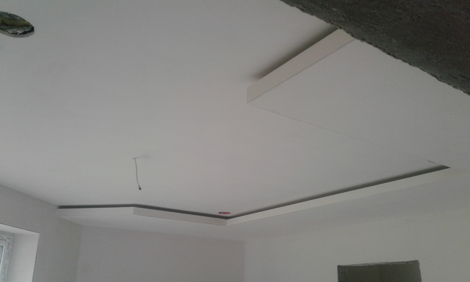 Biały sufit podwieszany z dekoracyjnymi listwami i otworem na oświetlenie, widoczne niedokończone wykończenia ścian.