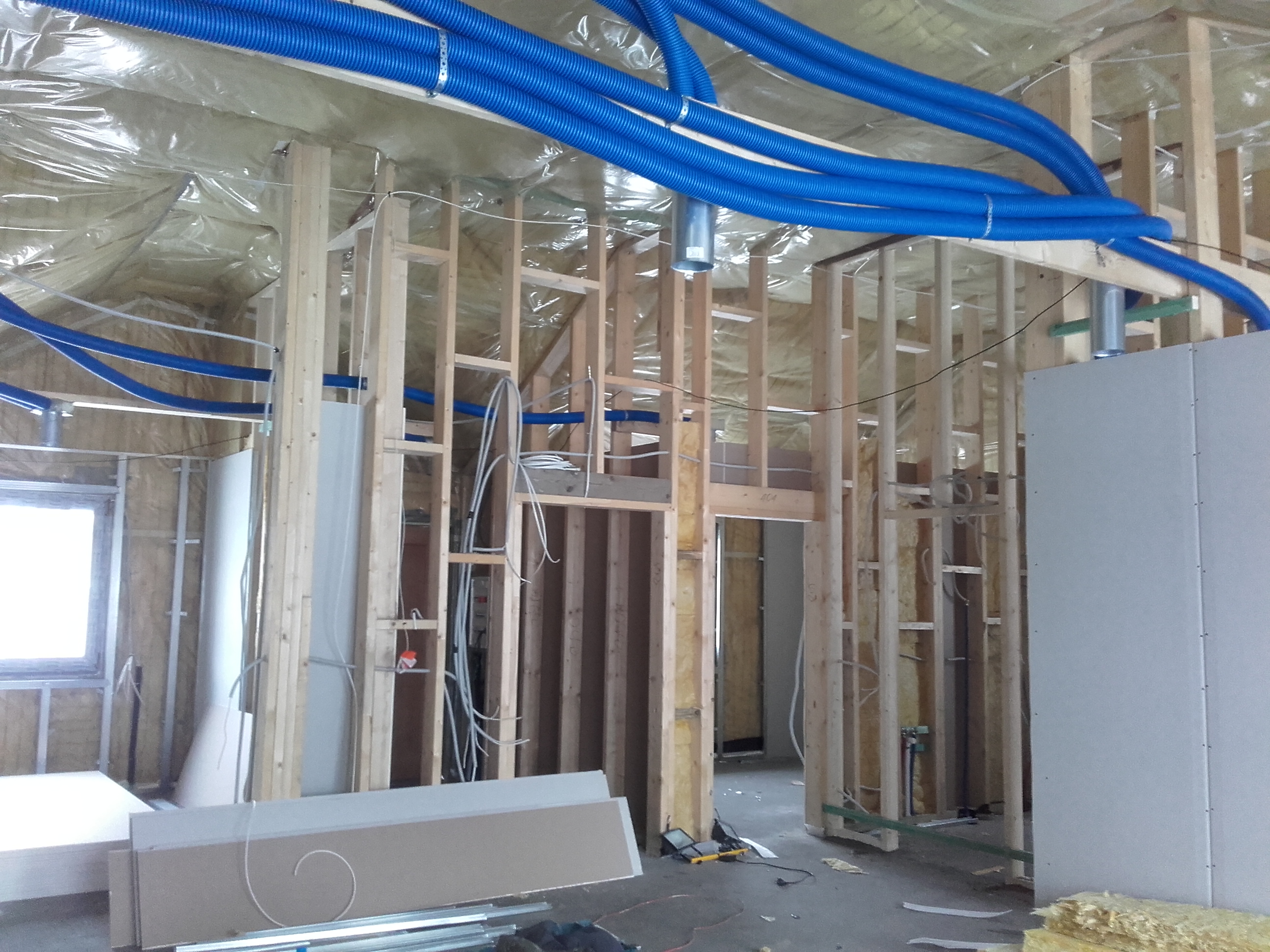 Wnętrze domu szkieletowego w trakcie budowy z widoczną konstrukcją drewnianą, izolacją folią, niebieskimi rurami wentylacyjnymi i płytami gipsowo-kartonowymi