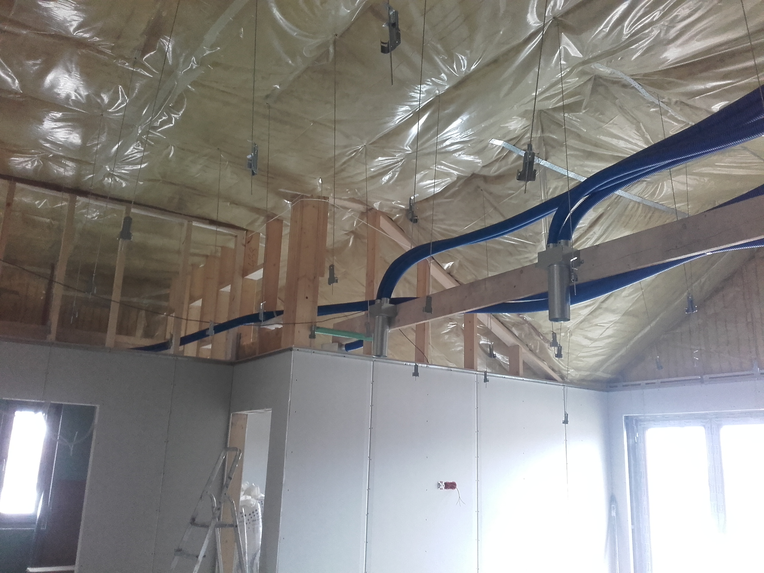 Wnętrze w trakcie budowy, widoczne drewniane elementy konstrukcyjne, folia izolacyjna na suficie, niebieskie rury wentylacyjne i ściany z płyt gipsowo-kartonowych z widoczną instalacją elektryczną...