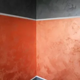 Pomarańczowo-brązowa ściana z efektem dekoracyjnym w rogu pomieszczenia, górna część ściany pomalowana na szaro, biała listwa sufitowa.