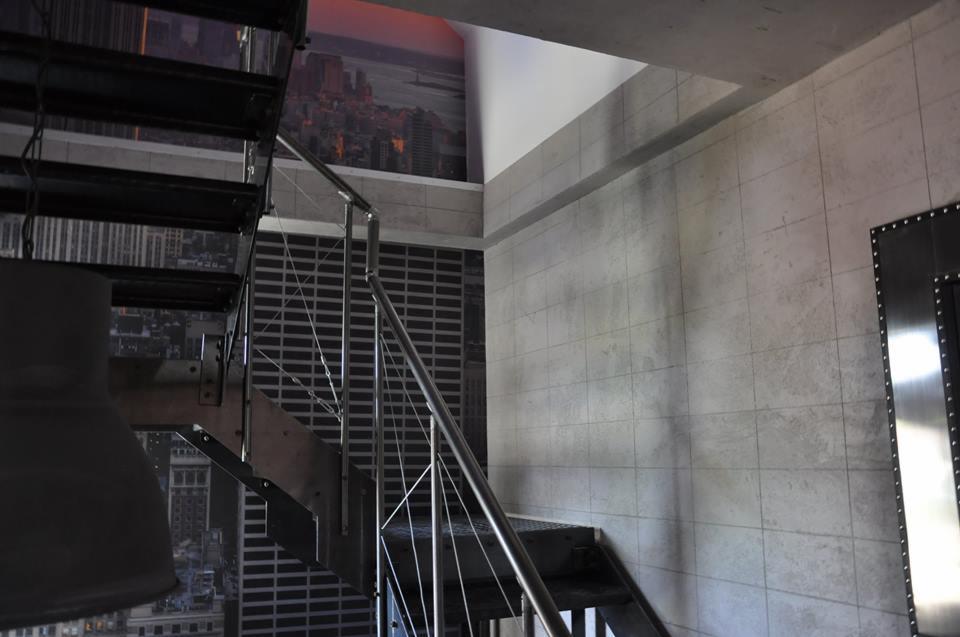 Metalowe schody z poręczą na tle ściany wykończonej płytkami imitującymi beton i fototapetą z panoramą miasta.