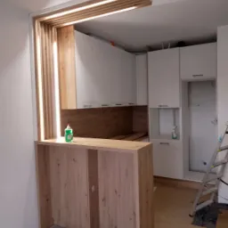 Wnętrze kuchni w trakcie remontu: drewniana zabudowa baru z oświetleniem, białe szafki kuchenne w tle, zabezpieczona podłoga i drabina. Widoczny etap prac wykończeniowych.