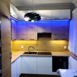 BUD-MER - Nowoczesna kuchnia w kształcie litery U z drewnianymi akcentami, białymi frontami i niebieskim oświetleniem LED. Czarny zlewozmywak i płyta indukcyjna na drewnianym blacie.