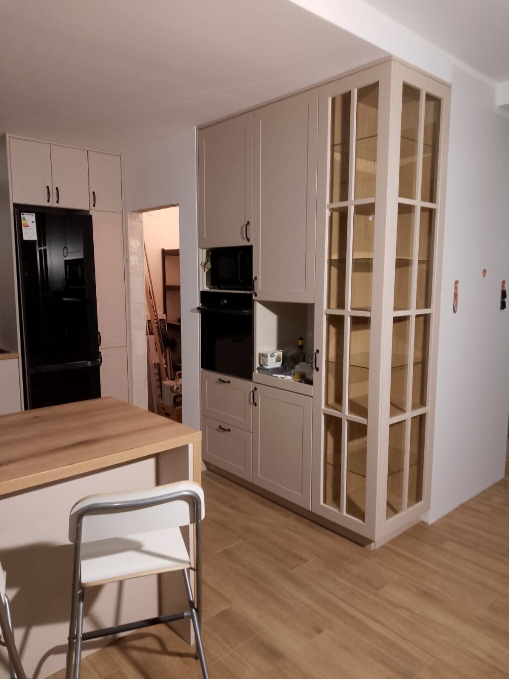 Jasna kuchnia z wyspą: zabudowa z szafkami, witryną i sprzętem AGD. Podłoga drewniana, krzesła barowe. Widok na otwarte przejście do innego pomieszczenia.