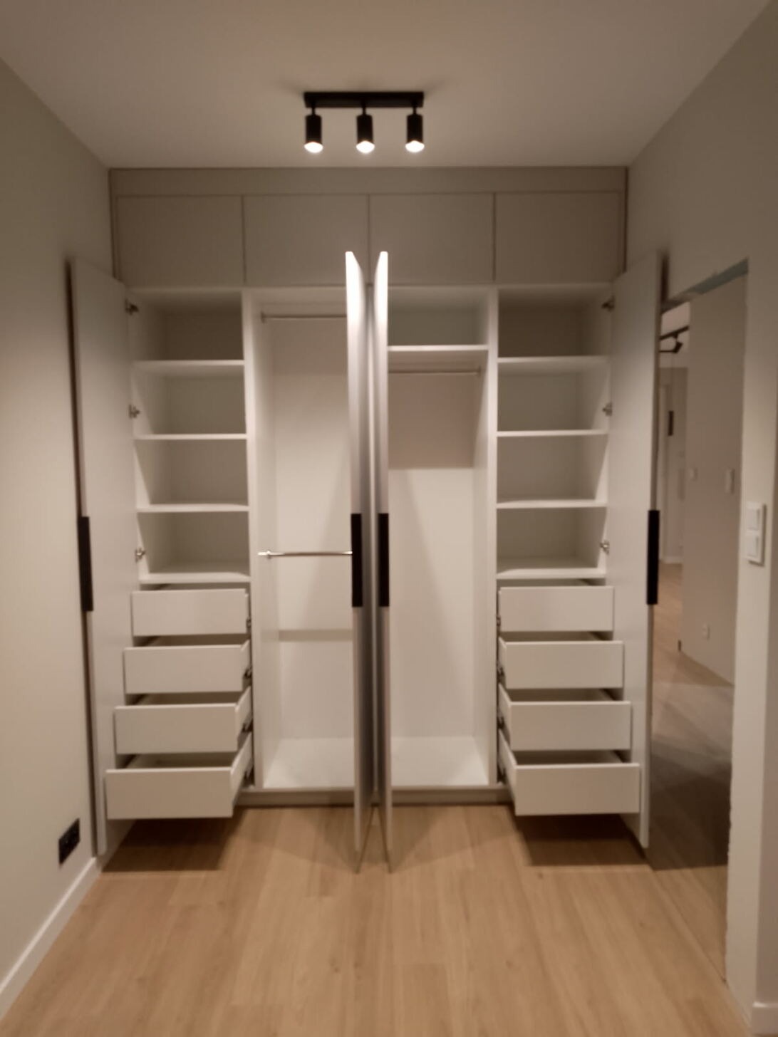 Jasny, nowoczesny, wnękowy system szaf z otwartymi drzwiami, ukazujący półki i szuflady w minimalistycznym wnętrzu z drewnianą podłogą i oświetleniem sufitowym.