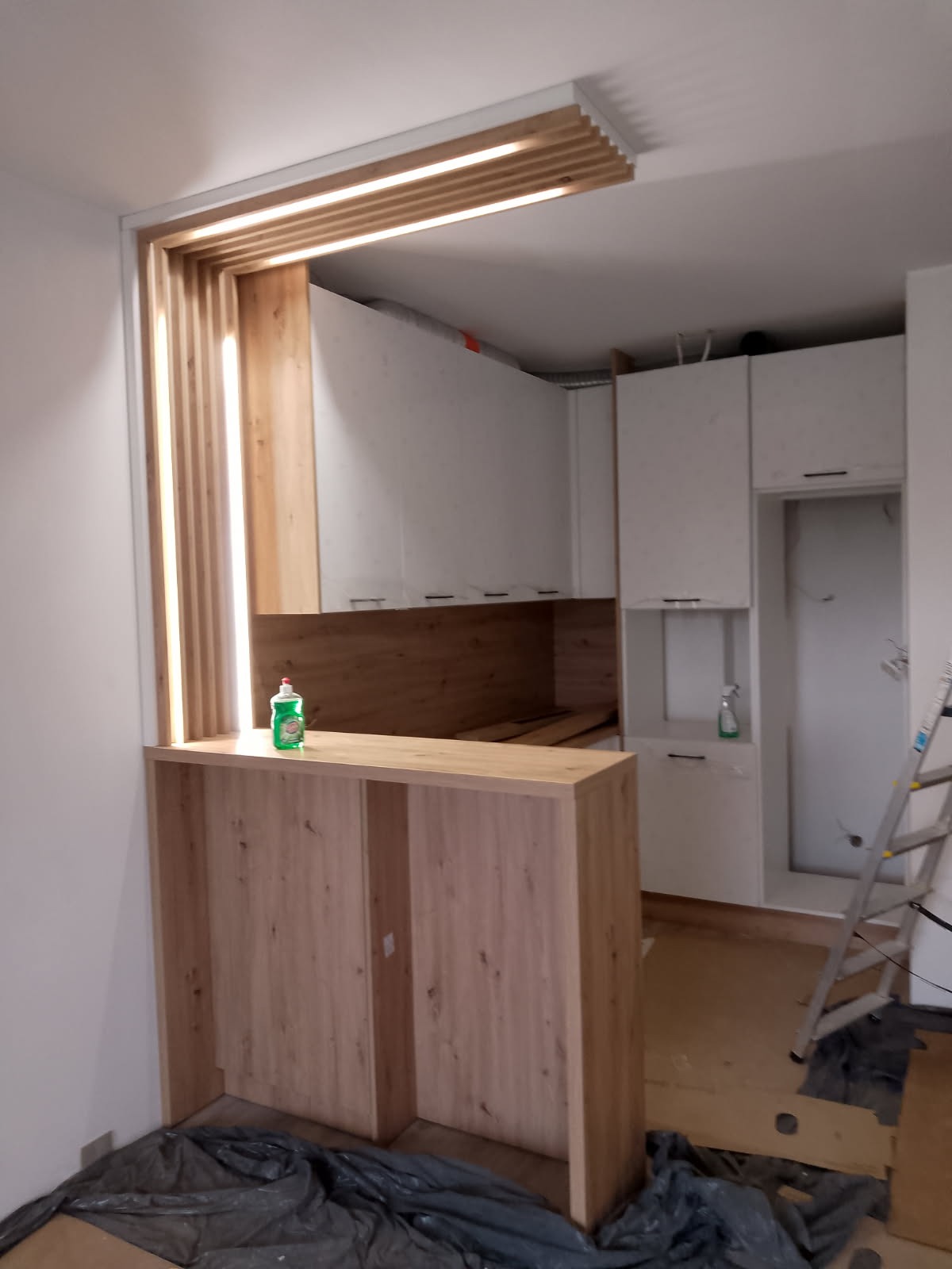 Wnętrze kuchni w trakcie remontu: drewniana zabudowa baru z oświetleniem, białe szafki kuchenne w tle, zabezpieczona podłoga i drabina. Widoczny etap prac wykończeniowych.