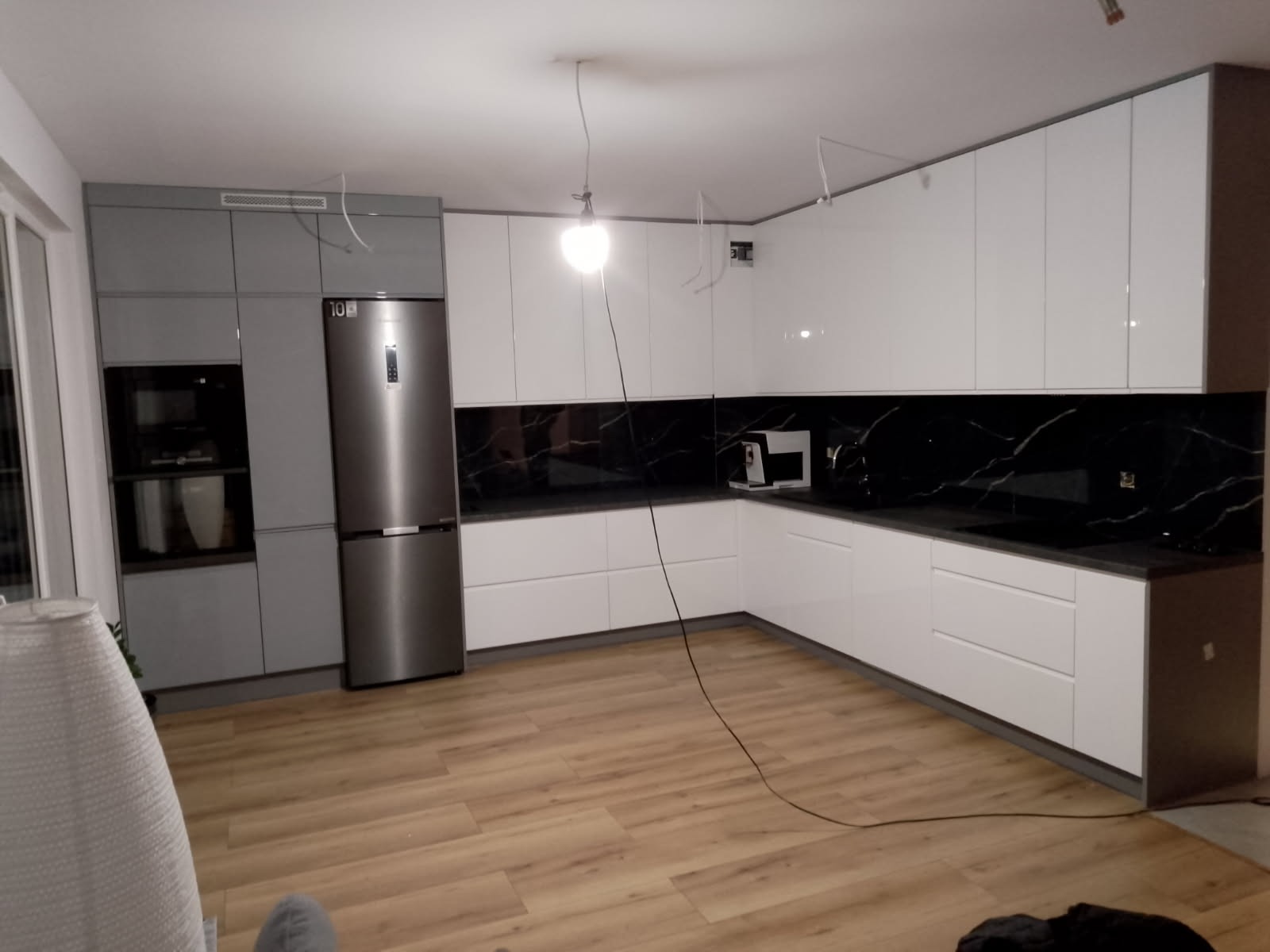Wykończona kuchnia z błyszczącymi białymi szafkami i czarnym blatem z marmurowym wzorem. Na podłodze panele imitujące drewno. Widoczne wiszące przewody.