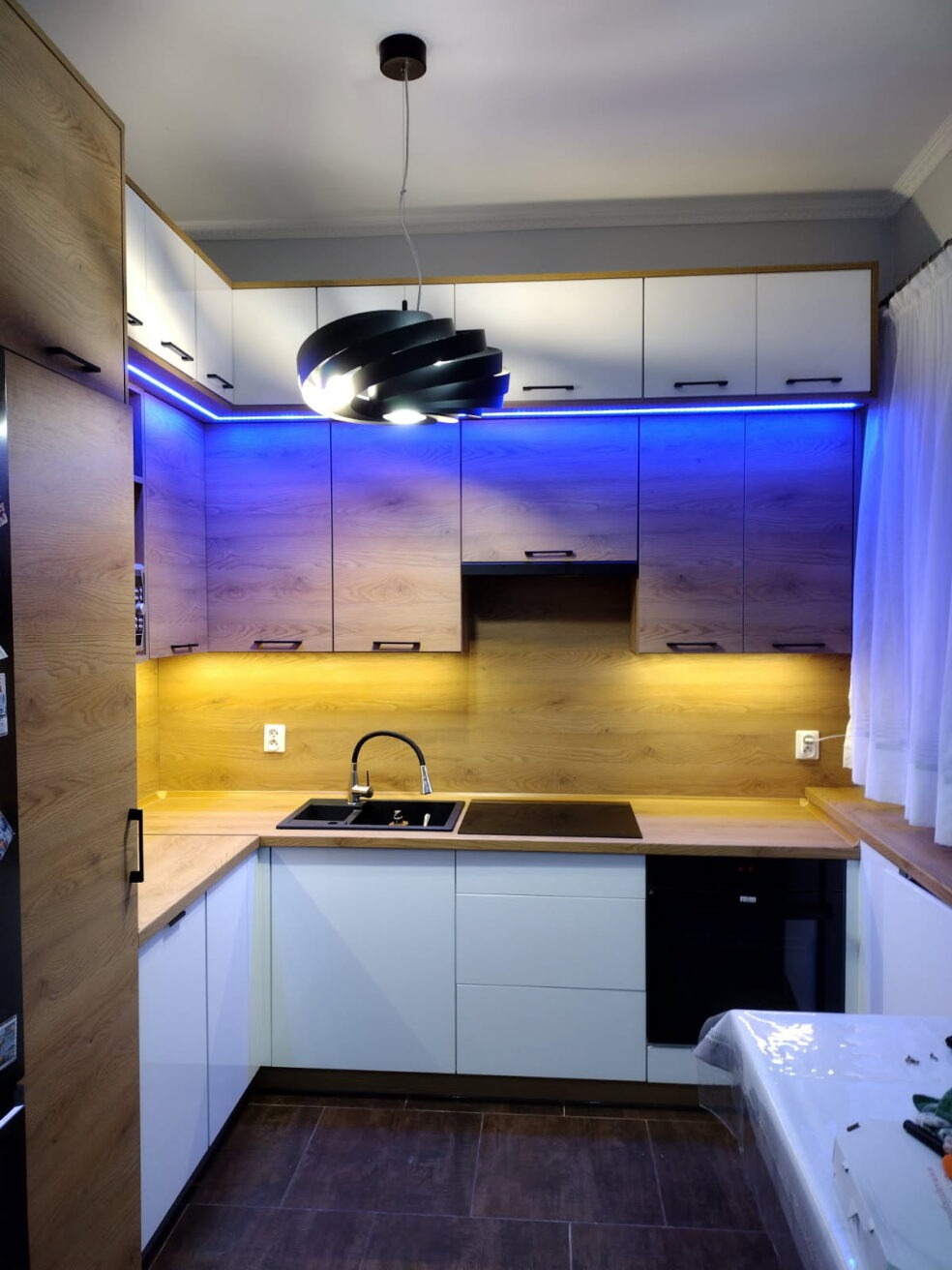 Nowoczesna kuchnia w kształcie litery U z drewnianymi akcentami, białymi frontami i niebieskim oświetleniem LED. Czarny zlewozmywak i płyta indukcyjna na drewnianym blacie.