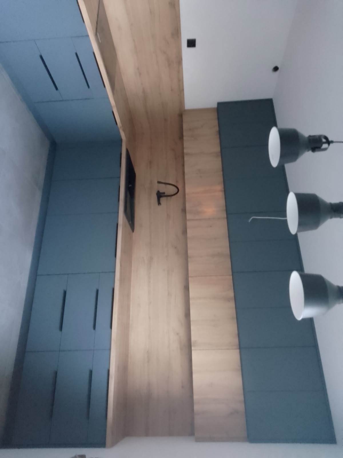 Nowoczesna kuchnia z niebieskimi szafkami i drewnianym blatem, oświetlona trzema lampami. Ujęcie z dołu podkreśla pionowe linie zabudowy.