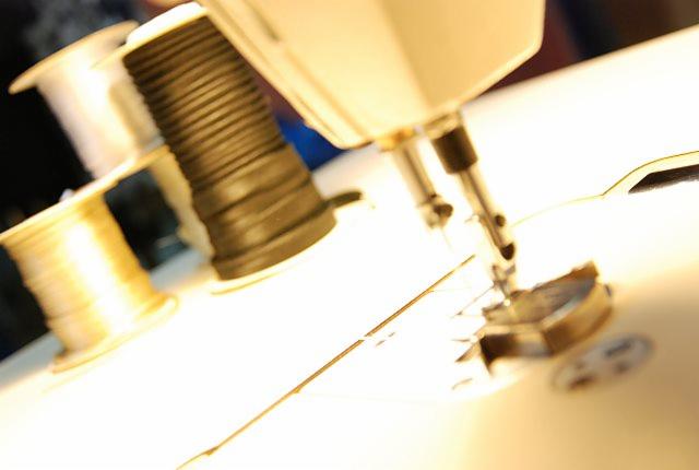 Maszyna do szycia w trakcie pracy, igła w ruchu, nici w kolorze ecru i czarna szpulka w tle. Ujęcie z bliska, oświetlenie studyjne.