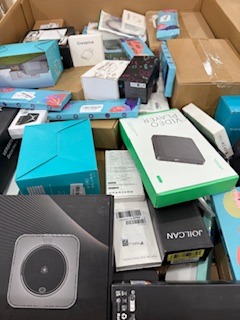 Stos pudełek z elektroniką i akcesoriami, widoczne różne marki i modele, chaotyczne ułożenie, jasne oświetlenie, pudełka częściowo zasłaniają się nawzajem.