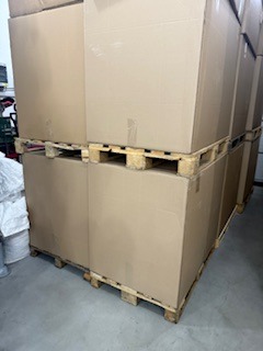 Stos kartonowych pudeł na drewnianych paletach, ustawionych jeden na drugim w magazynie. Widoczne ściany i podłoga magazynu, oświetlenie górne.