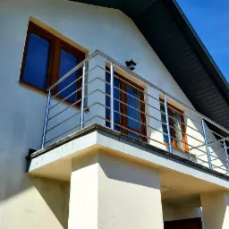 Balkon z nowoczesną, metalową barierką z rurek, widok z dołu na tle błękitnego nieba.