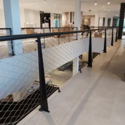 Metalowa siatka zabezpieczająca schody w nowoczesnym wnętrzu, czarne słupki podtrzymujące, widoczne osoby w tle.