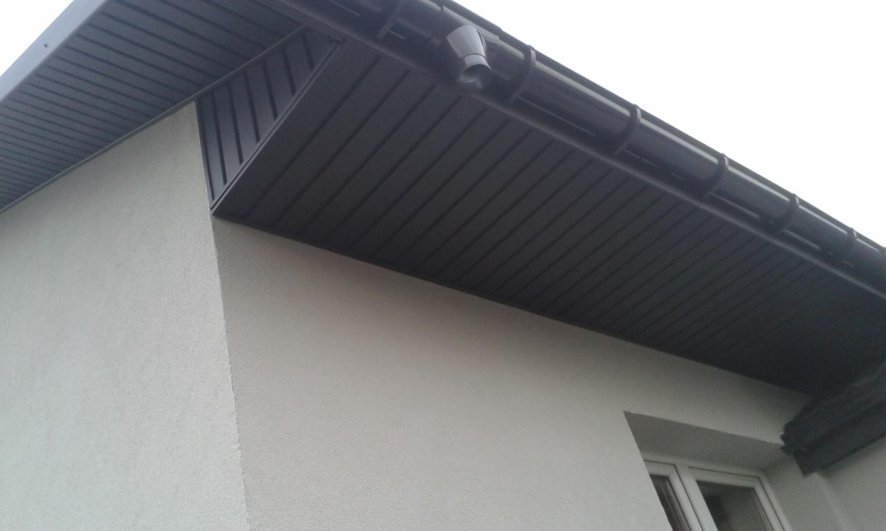 Fragment elewacji domu z ciemną, podbitką dachową z widoczną rynną i rurą spustową.
