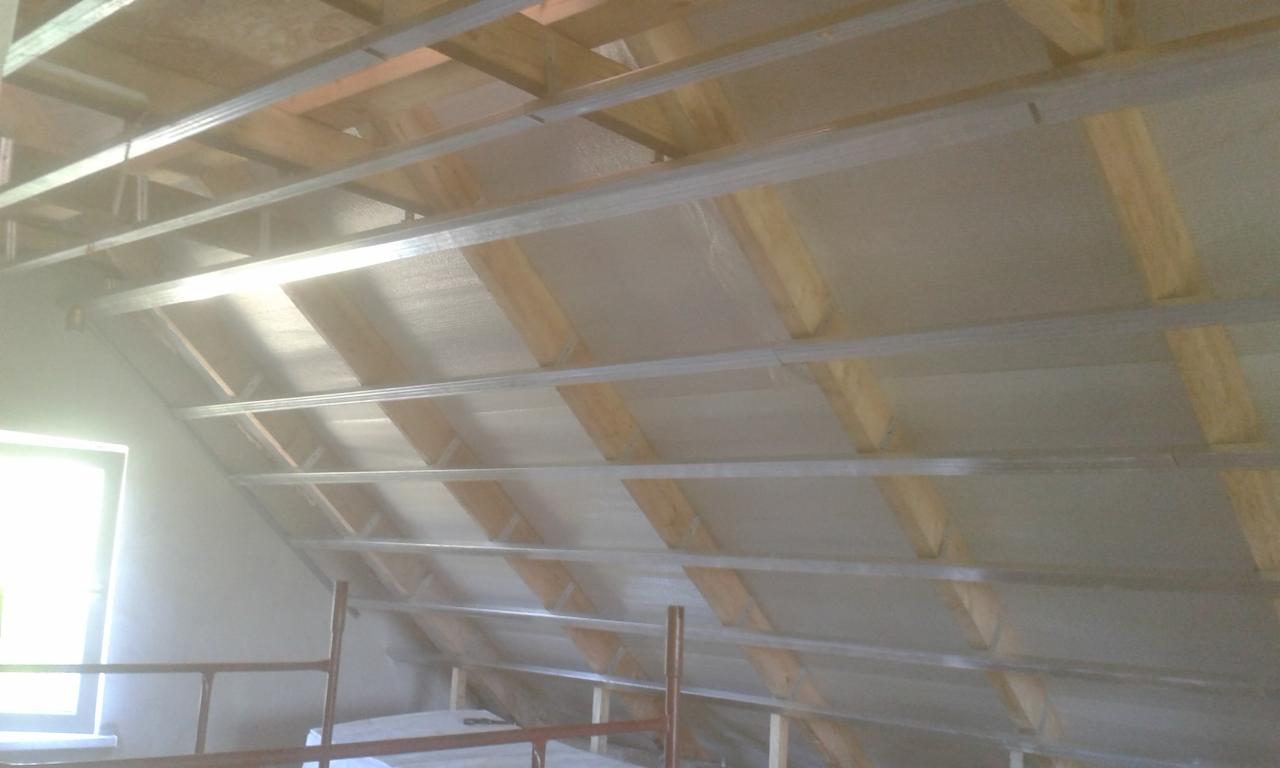 Skośny strop poddasza w trakcie montażu, widoczne drewniane belki, metalowe profile i płyty pokrywające izolację, okno w tle.