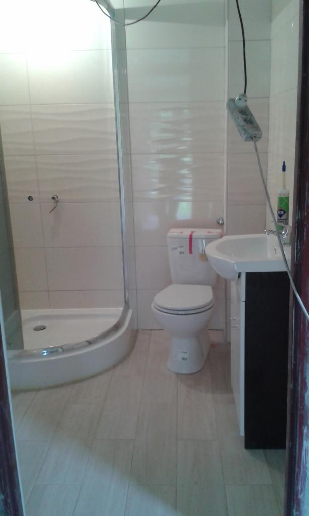 Niedokończona łazienka z białą kabiną prysznicową, toaletą i umywalką z szafką, widoczne kable i przedłużacz, płytki na ścianach i podłodze.