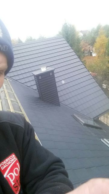 Nowy, grafitowy dach z kominem i oknami połaciowymi, widok z góry, osoba w bluzie z logo firmy dekarskiej w kadrze.