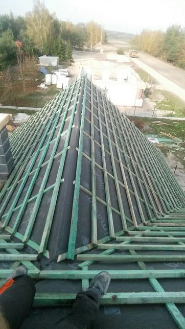 Wysoki dach w trakcie budowy, widok z góry na konstrukcję z łat i kontrłat pokrytą membraną dachową, w tle krajobraz z budynkiem w stanie surowym otwartym.