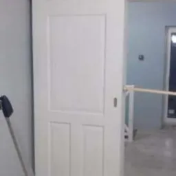 Białe, przesuwne drzwi wewnętrzne z trzema panelami, zamontowane w jasnym pomieszczeniu. Widoczny fragment drabiny i cegły na podłodze.