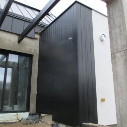 Ecofasada PRO - Nowoczesna elewacja domu z czarną drewnianą okładziną i białym tynkiem, widoczny fragment pergoli i okna. Surowy betonowy element konstrukcyjny w prawym rogu.