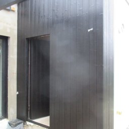 Ecofasada PRO - Ciemna, drewniana elewacja budynku z widocznym wejściem i fragmentem okna. Świeżo pomalowane deski, surowy stan otoczenia. Detal wykończenia.