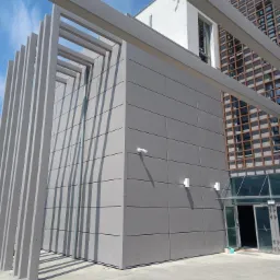 Nowoczesny budynek z szarą elewacją panelową, widoczne elementy konstrukcyjne pergoli i fragmenty instalacji elektrycznej na posadzce.