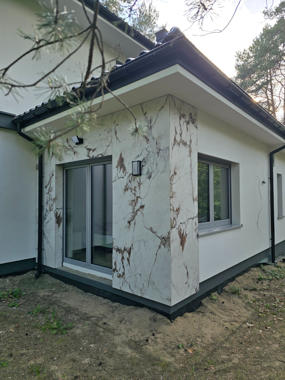 Wykończona elewacja domu z imitacją marmuru, widoczne okna i lampa zewnętrzna. Detal narożnika z listwą przypodłogową. W tle drzewa i fragment gałęzi sosny.