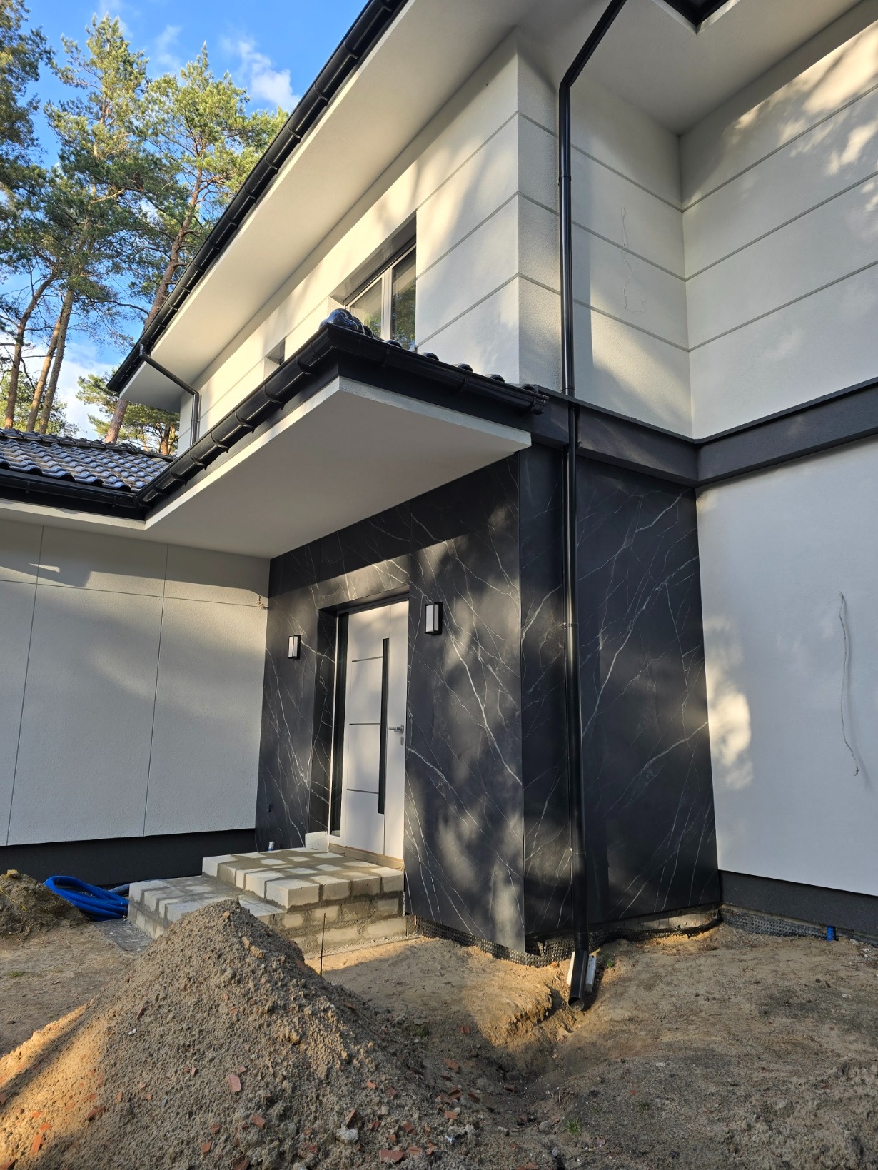 Nowoczesny dom z elewacją w odcieniach bieli i czerni, wykończony marmurkowym wzorem przy wejściu. Widoczne schody wejściowe i fragment ogrodu z ziemią.