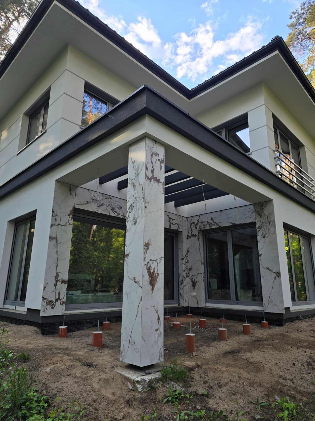 Nowoczesny dom z elewacją imitującą marmur i zadaszeniem tarasu wspartym na kolumnach. Widoczne przygotowania do zagospodarowania terenu wokół budynku.