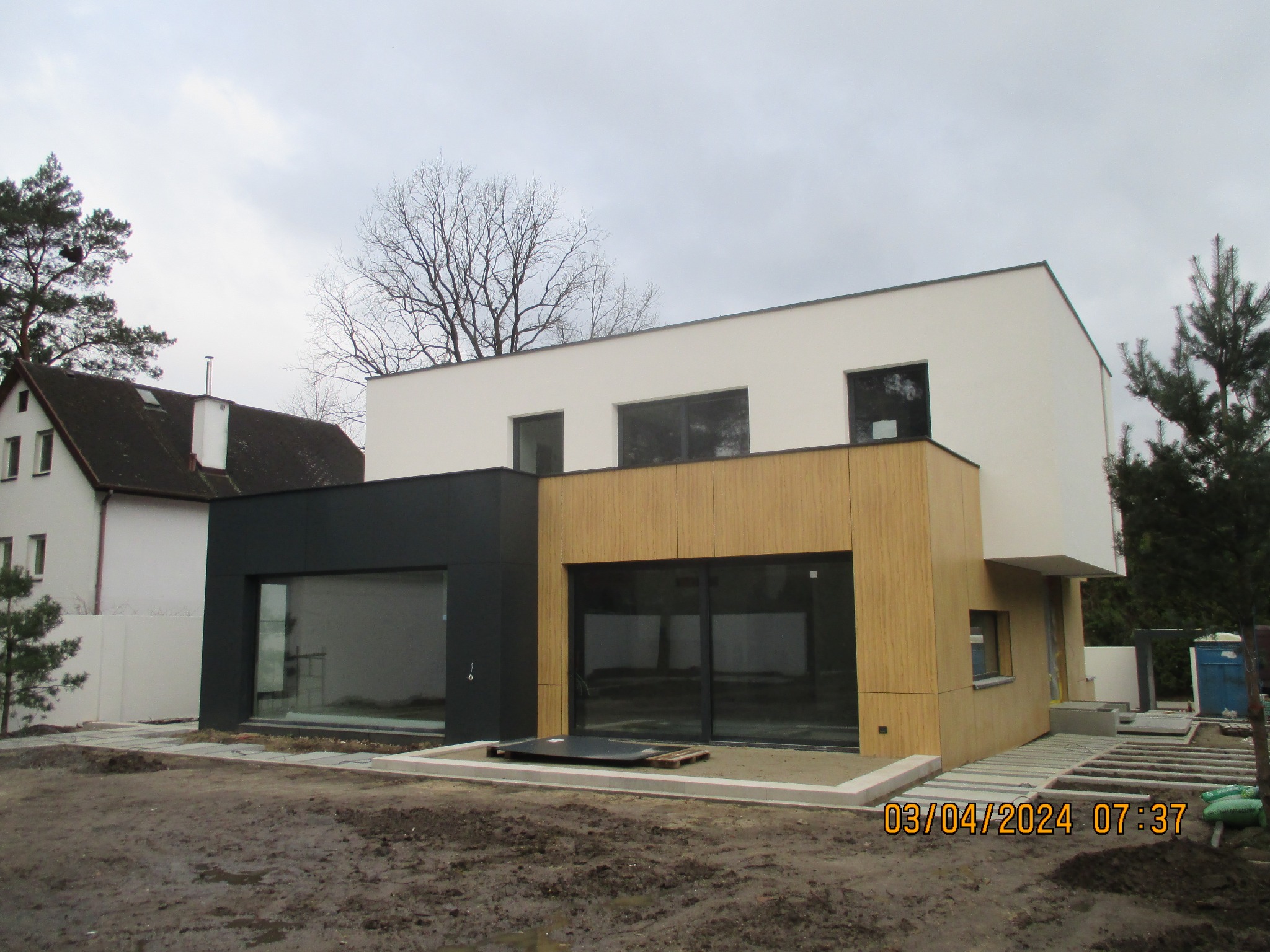 Nowoczesny dom z elewacją z drewna i czarnymi panelami. Widoczne okna i fragment tarasu w trakcie budowy. Pochmurne niebo w tle.