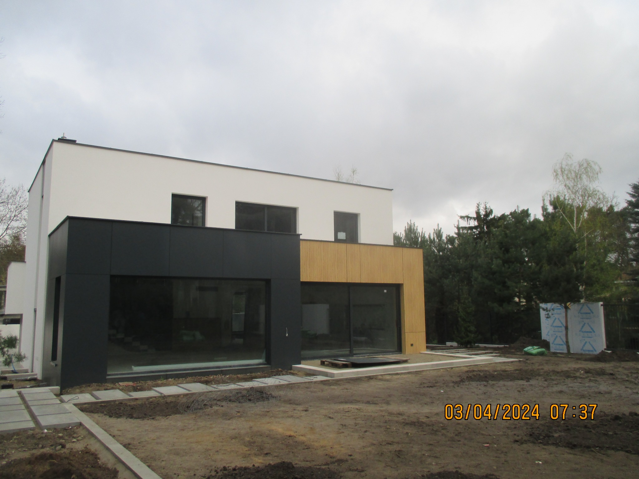 Nowoczesny dom jednorodzinny z elewacją w odcieniach bieli, czerni i drewna, duże okna, Warszawa. Widok z zewnątrz, teren wokół domu w trakcie prac wykończeniowych.