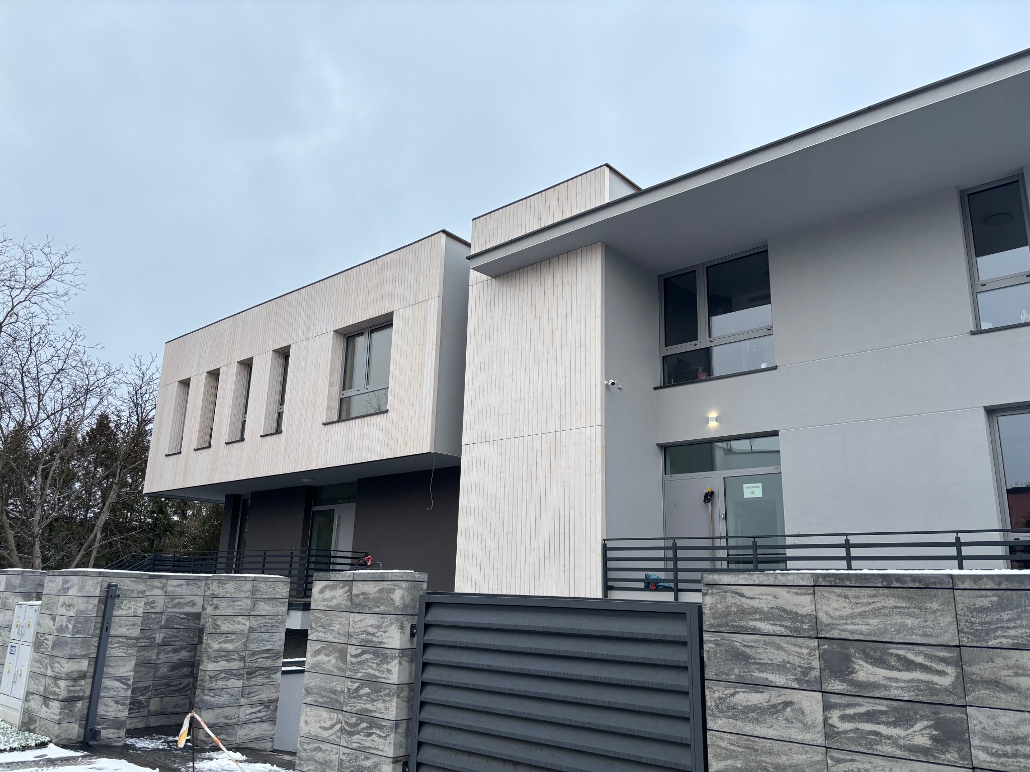 Nowoczesny dom z jasną elewacją z pionowych desek i szarą, gładką fasadą. Widoczny fragment ogrodzenia z szarego kamienia. Pochmurne niebo w tle.