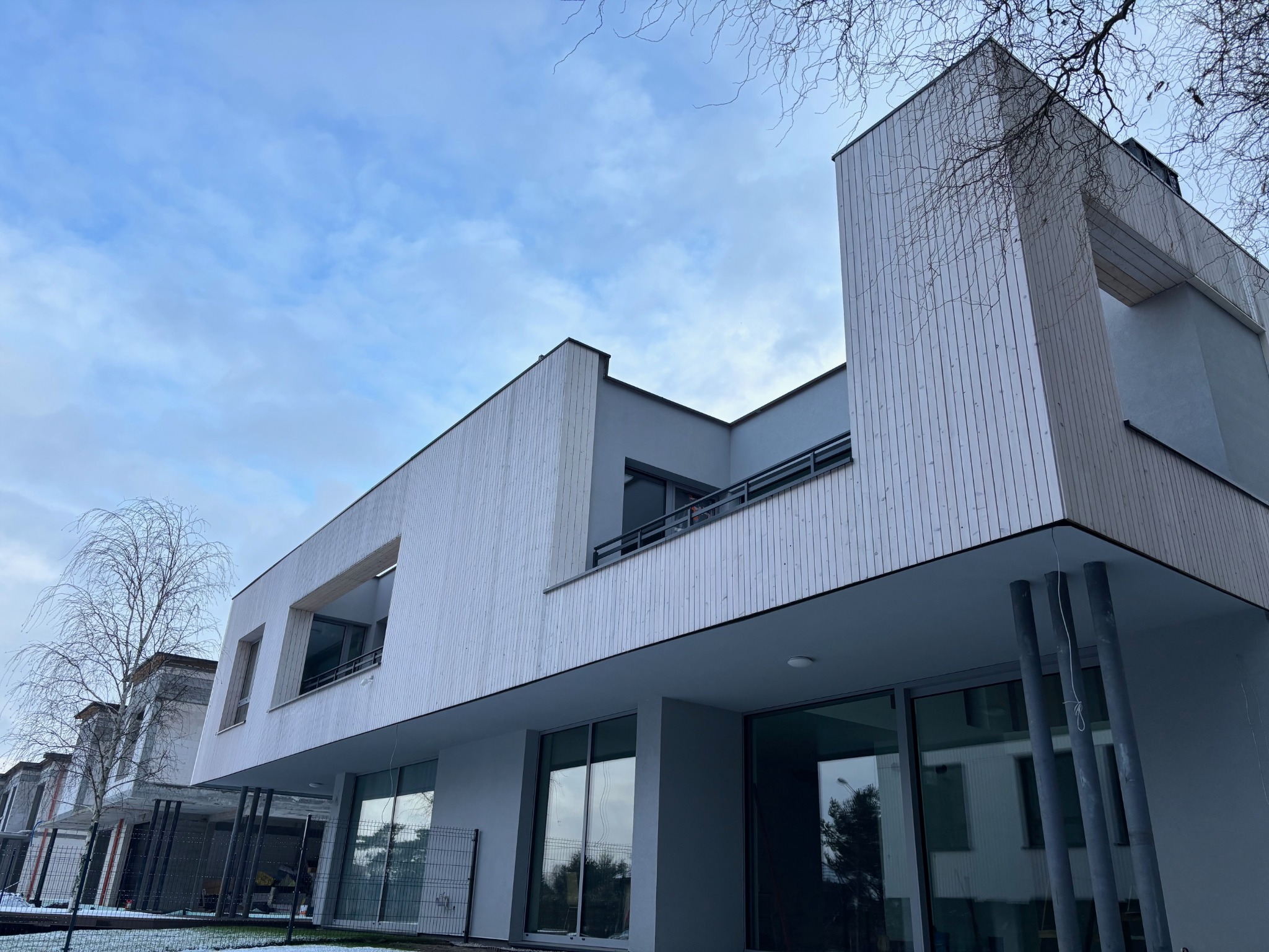 Nowoczesny dom z jasną, drewnianą elewacją w pionowe pasy, widoczny z dołu pod kątem. Minimalistyczny design, duże okna, szare elementy wykończeniowe i błękitne niebo w tle.