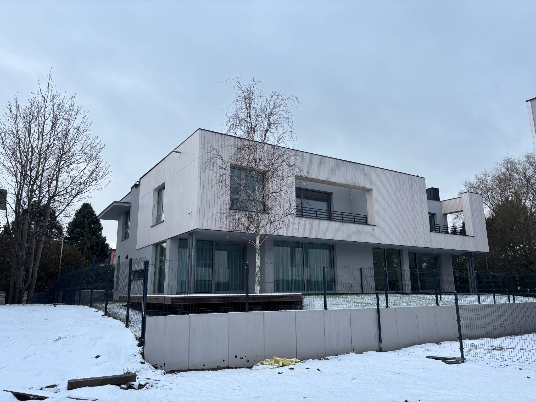 Nowoczesny dom z elewacją z jasnego drewna, widoczny zimą. Minimalistyczny design, duże okna, balkony. W tle drzewa i pochmurne niebo.