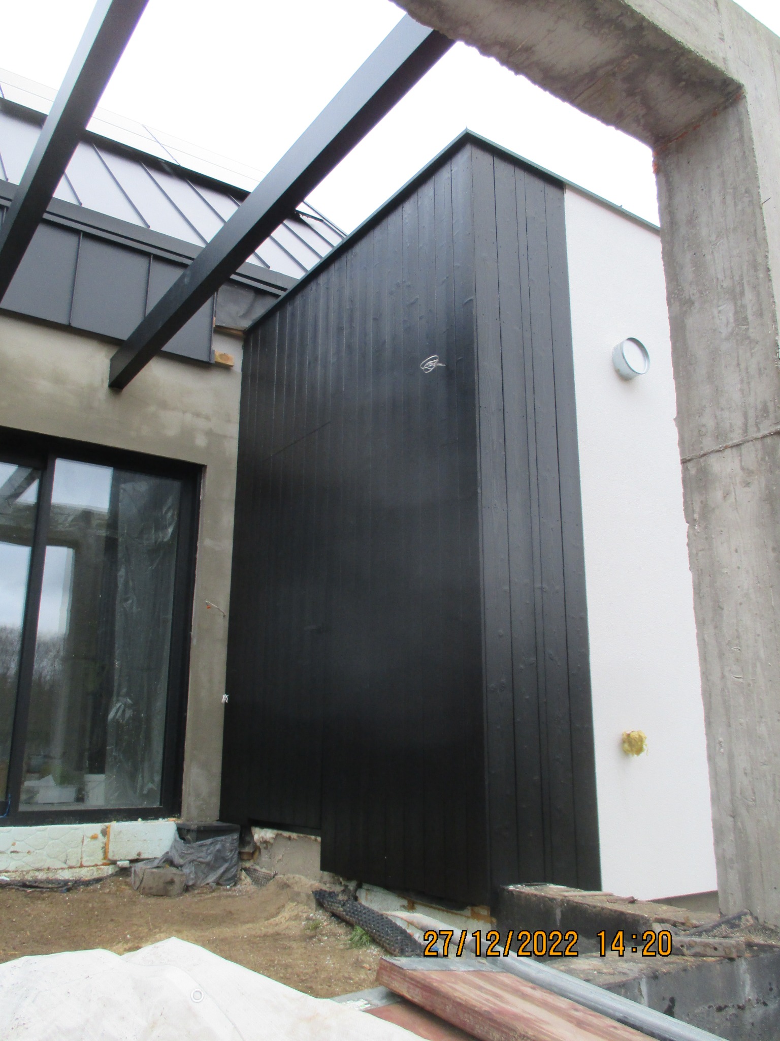 Nowoczesna elewacja domu z czarną drewnianą okładziną i białym tynkiem, widoczny fragment pergoli i okna. Surowy betonowy element konstrukcyjny w prawym rogu.