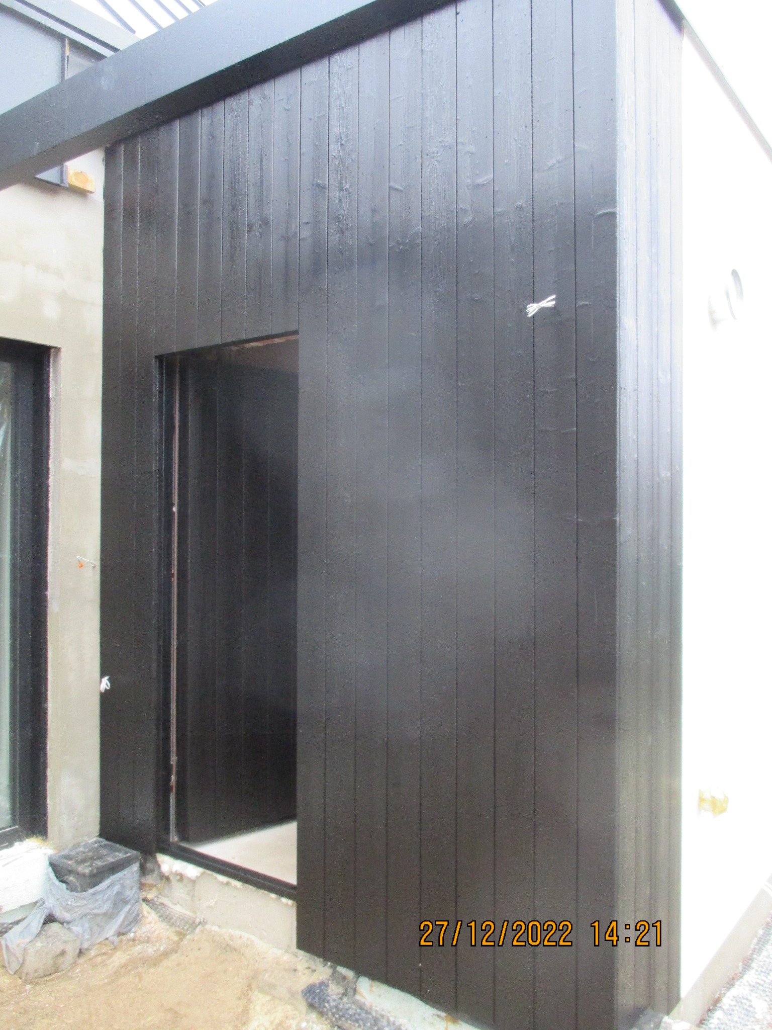 Ciemna, drewniana elewacja budynku z widocznym wejściem i fragmentem okna. Świeżo pomalowane deski, surowy stan otoczenia. Detal wykończenia.