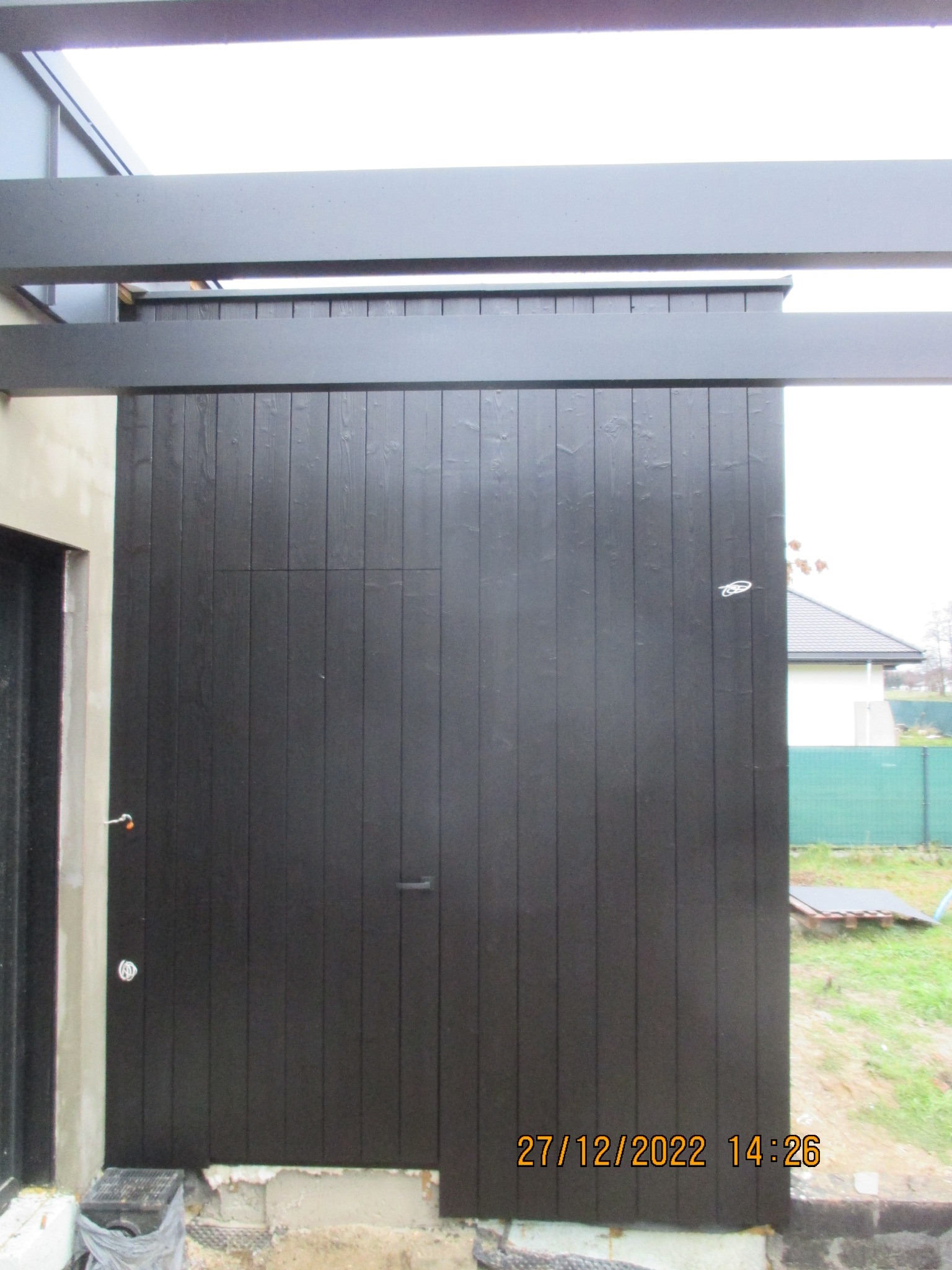 Ciemna, drewniana ściana z ukrytymi drzwiami, fragment pergoli z czarnych belek, surowy stan wykończenia przy gruncie. Detal nowoczesnej architektury.