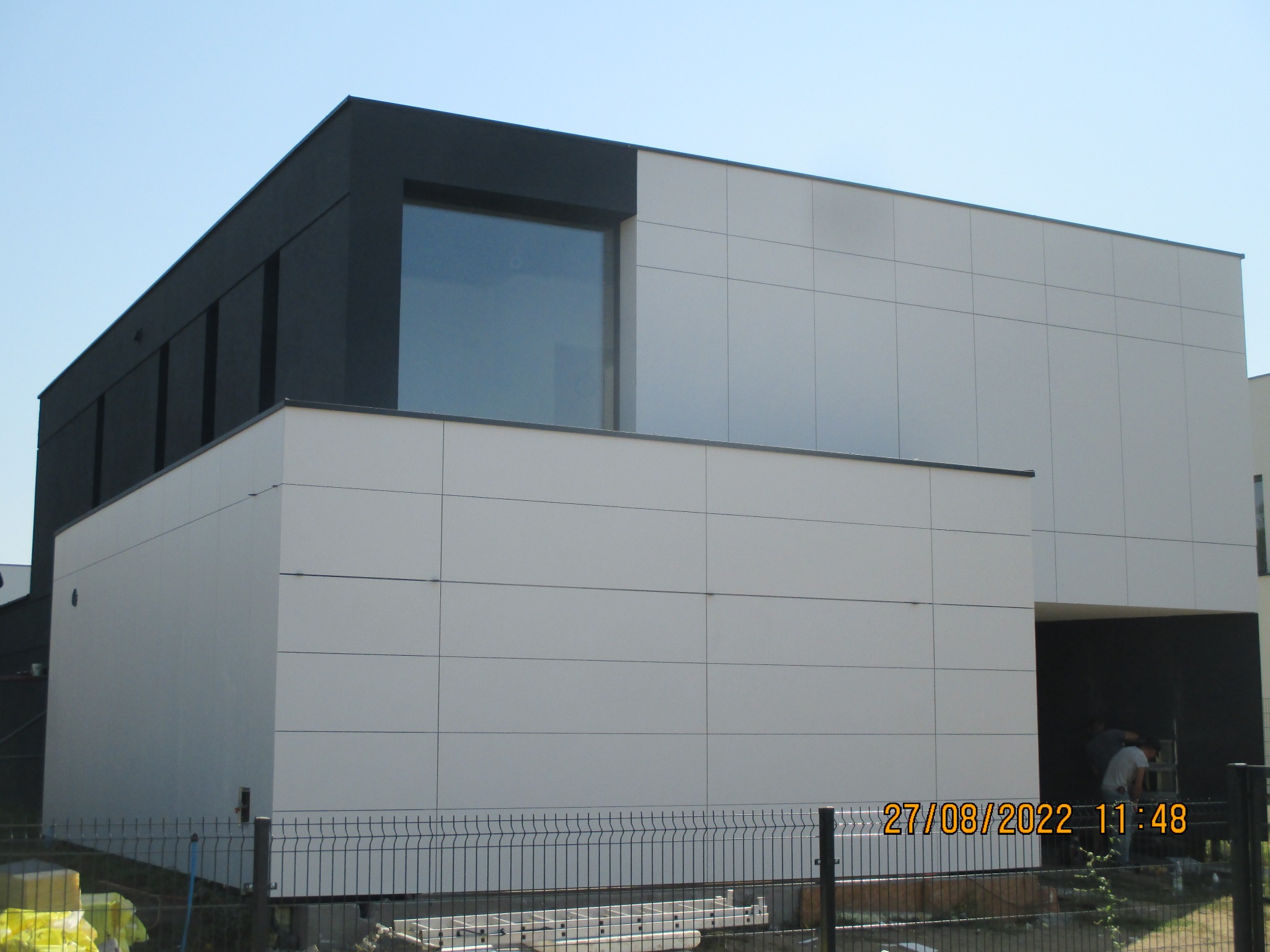 Nowoczesny budynek z elewacją z paneli w kolorze białym i czarnym, widoczna drabina oparta o budynek, zarys sylwetek osób w przejściu, ogrodzenie siatkowe na pierwszym planie.